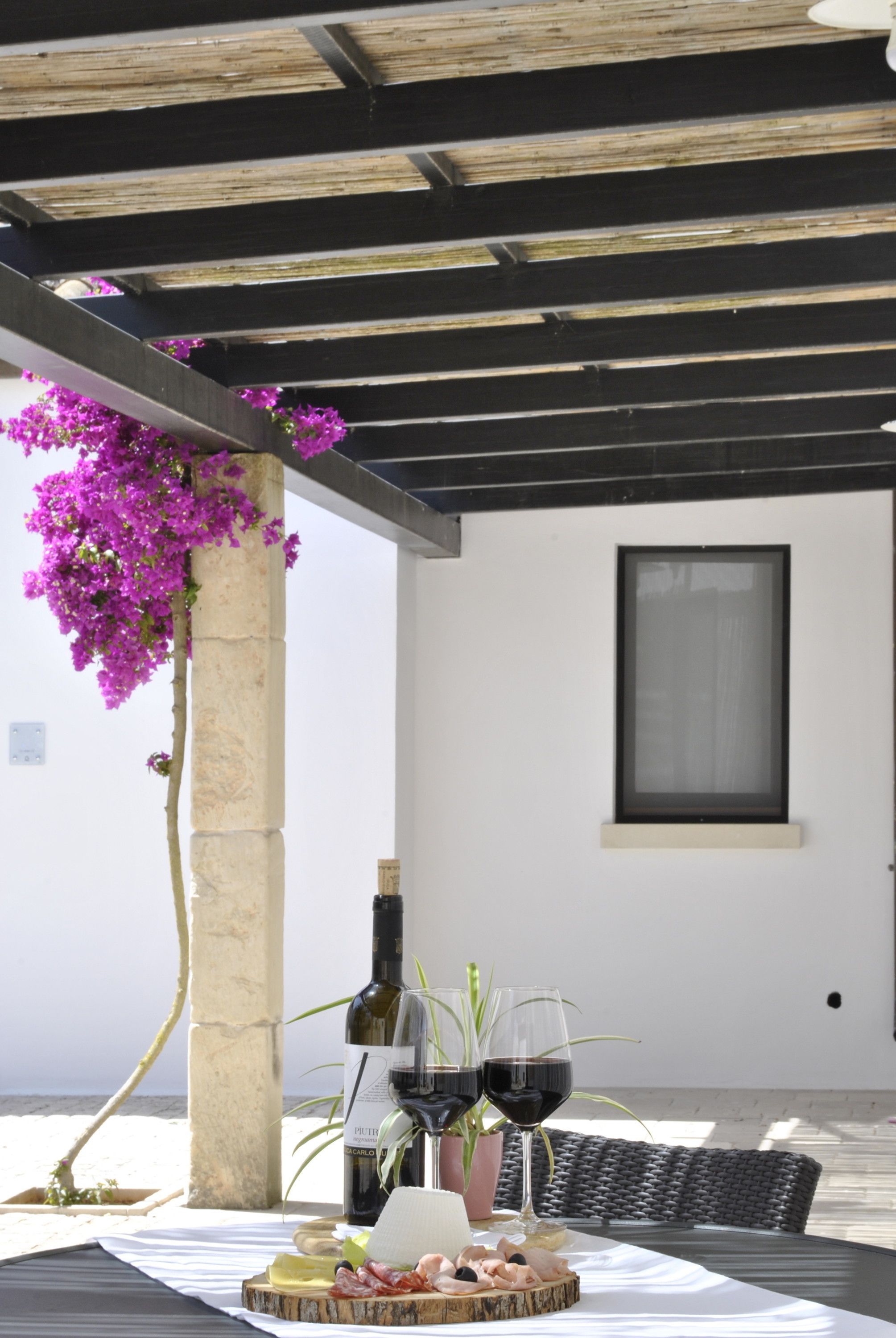 A covered outdoor dining area with a table set for two, featuring a bottle of wine, two glasses, a small plant, and a plate of food.