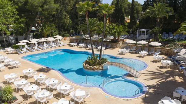 Zona de piscina en un resort con múltiples tumbonas, una piscina central y vegetación circundante.