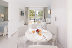 A modern hotel room with a dining area, a balcony with a view, and a bed with a blue bedspread.