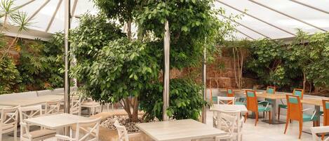 Una zona d'exterior per dinar amb diverses taules i cadires, envoltada de vegetació abundant i una estructura de sostre de vidre.