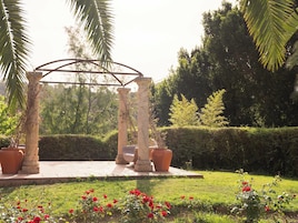 Un jardín con un pérgola de piedra, plantas en macetas y un césped con flores rojas.