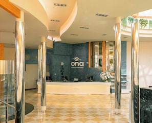 A reception area with a check-in counter, a logo for 'ona Hotels & Apartments', and a modern interior design with checkered flooring and metallic columns. - Ona Palmira Paguera (Calvia)