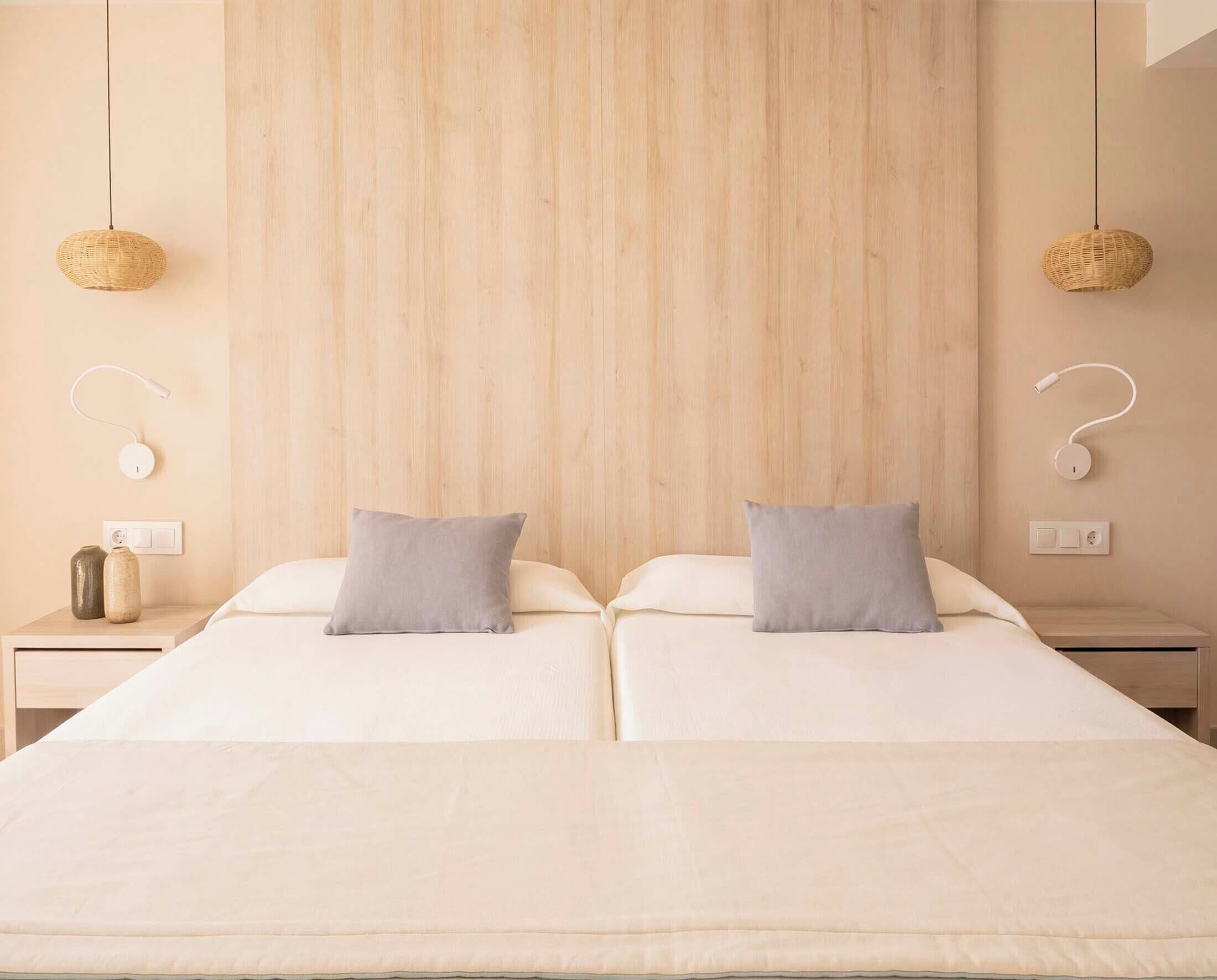 Cama doble con dos almohadas, mesita de noche con lámpara y enchufe en la pared.