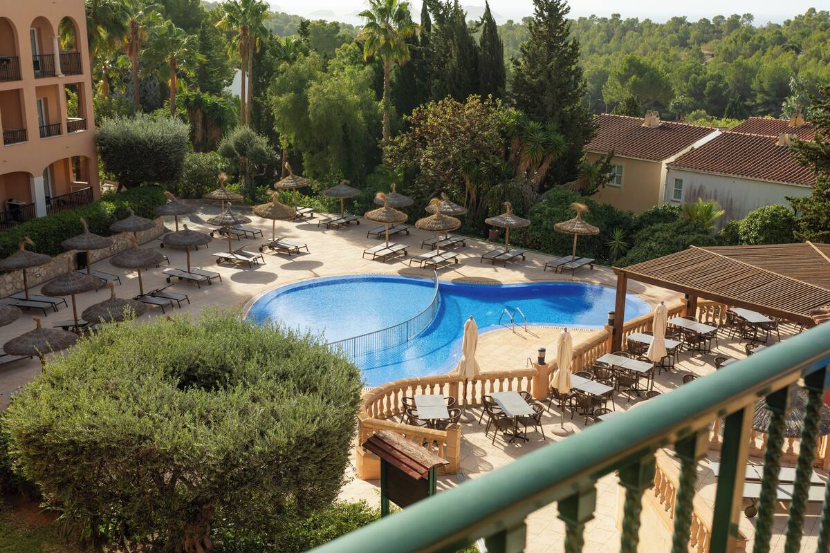 Un espace piscine aménagé dans un complexe hôtelier, avec des chaises longues et des parasols, entouré d’une végétation luxuriante et bordé d’un bâtiment dont le toit est recouvert de tuiles.