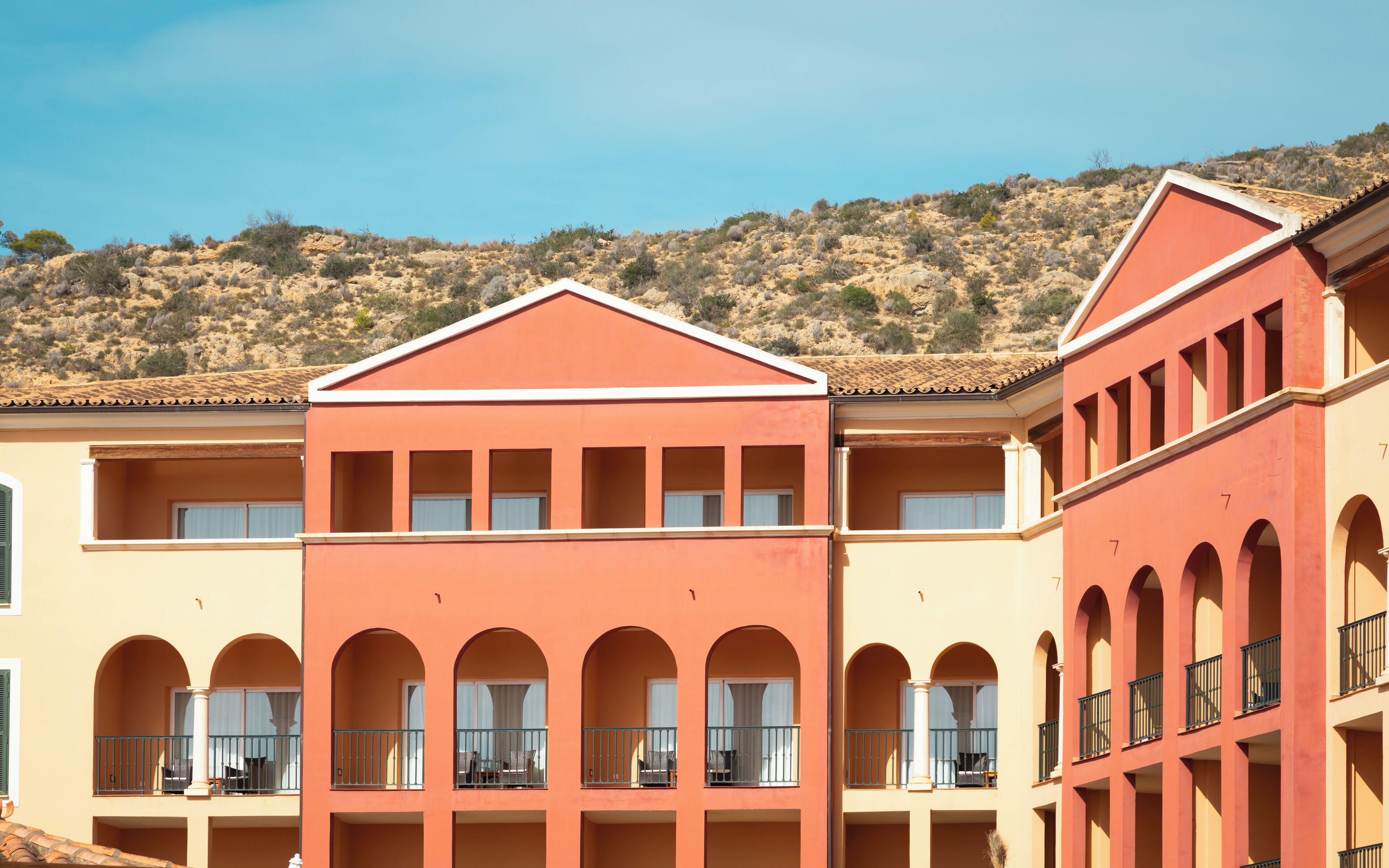 A multi-story hotel with a red facade, multiple balconies, and a hillside backdrop.