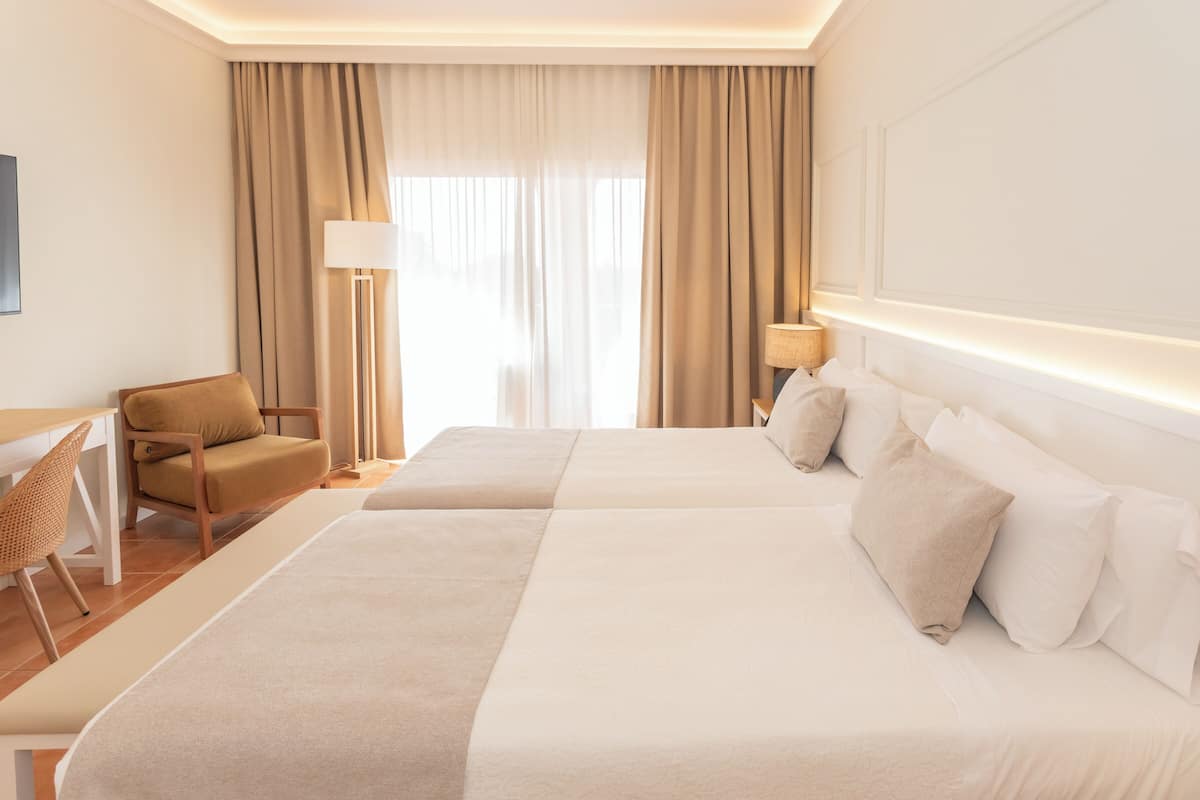 Une chambre d’hôtel avec deux lits, un bureau, une chaise et une grande fenêtre avec des rideaux.