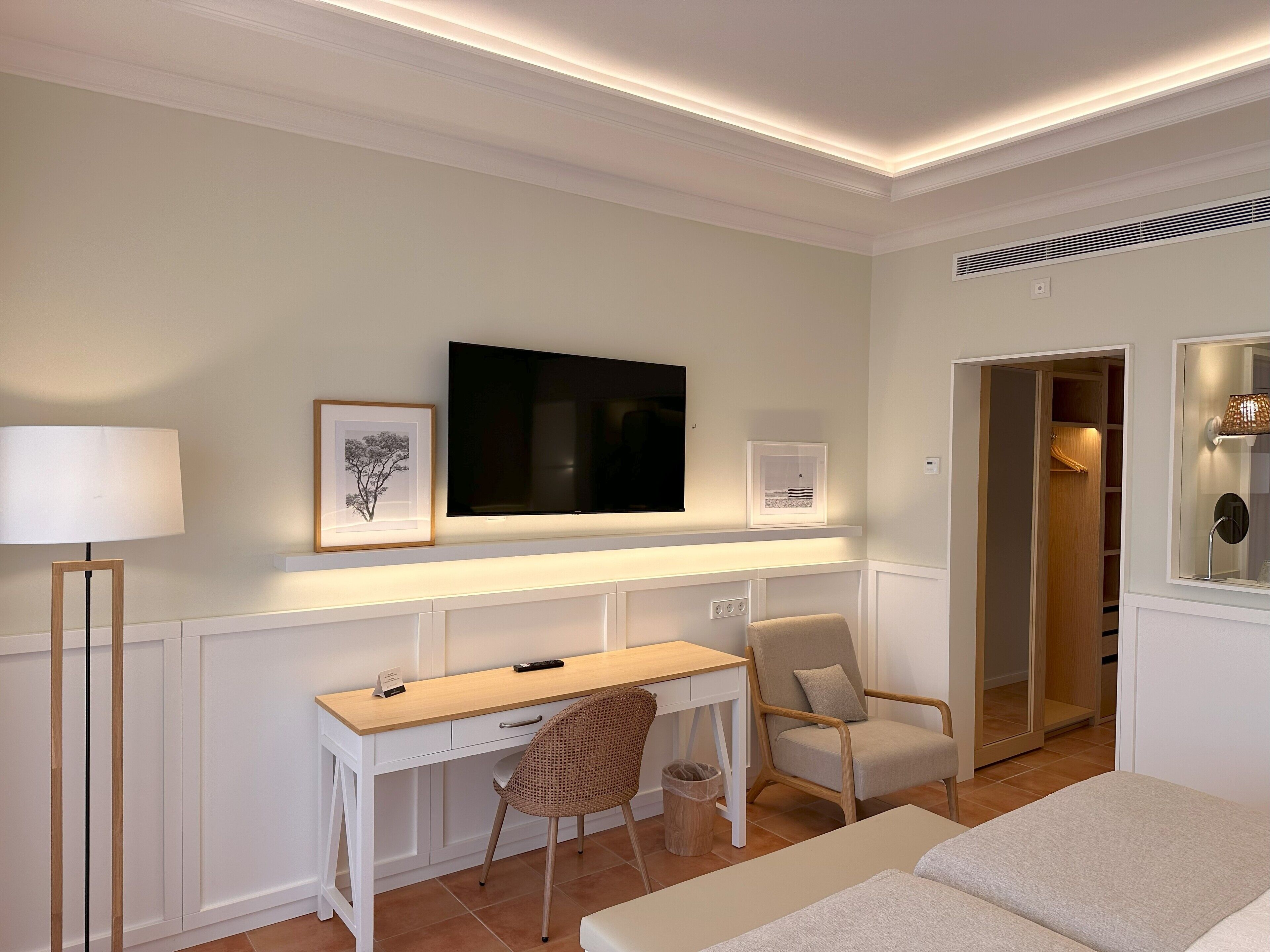 A hotel room with a white desk, a chair, a bed, a TV mounted on the wall, and a walk-in closet.
