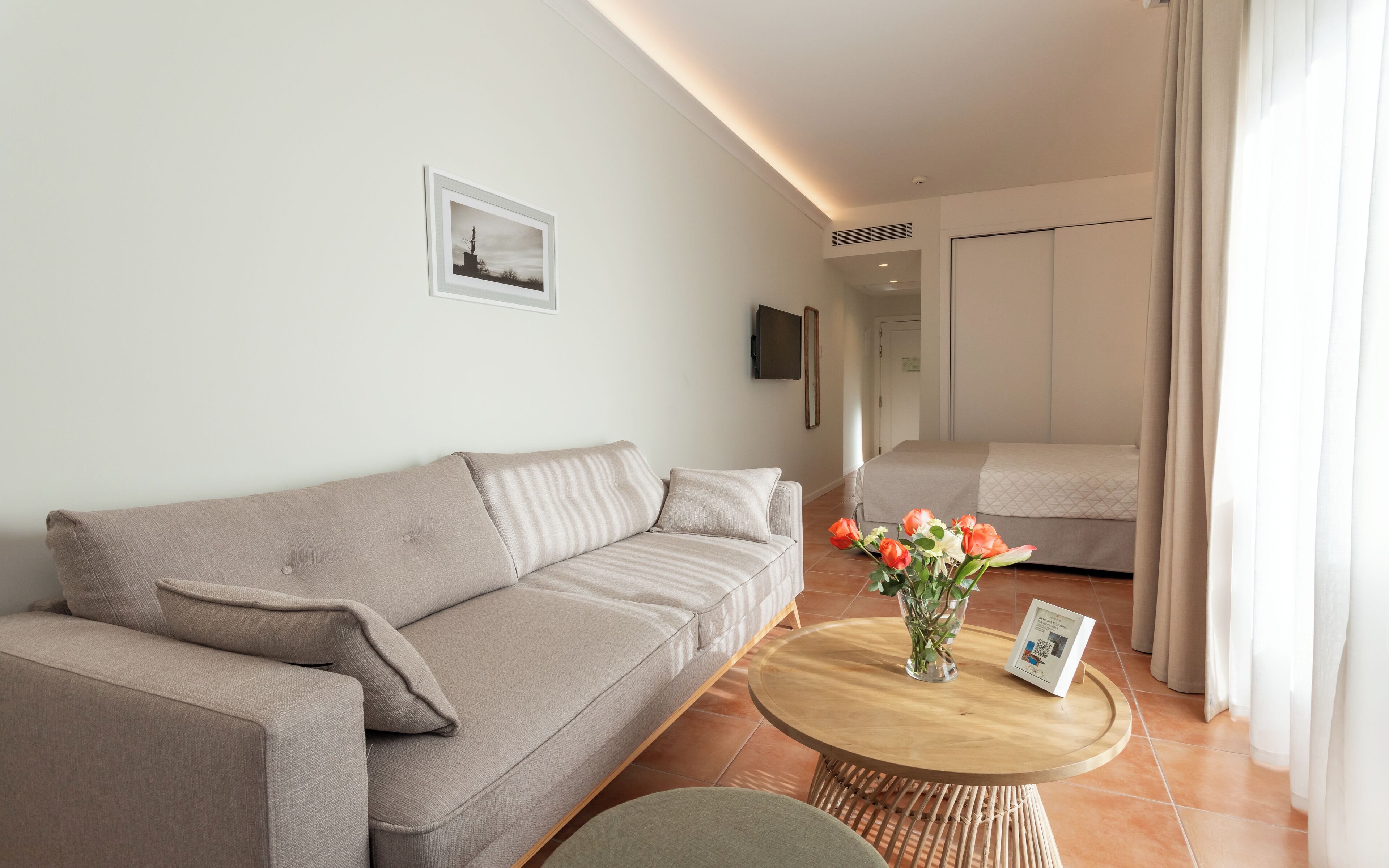 A modern living room with a sofa, a round coffee table with a vase of flowers, and a bed in the background.