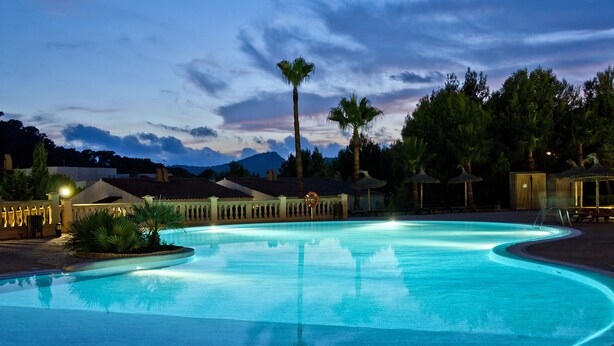 A large, clear swimming pool with a curved edge, surrounded by palm trees and a building with a tiled roof. - Hotel Bordoy Don Antonio (Calvia)