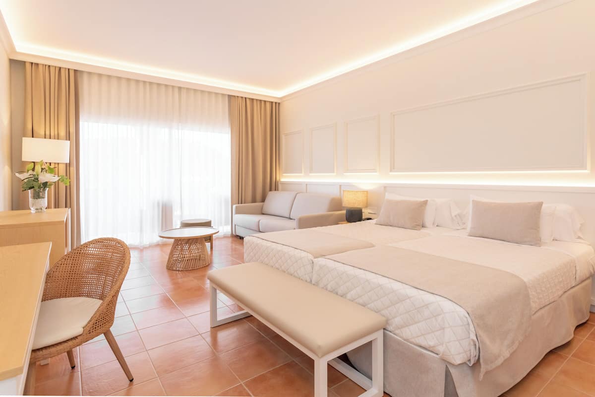 Une chambre d’hôtel avec un grand lit, un canapé, une petite table, un banc et un bureau avec une lampe et des fleurs.