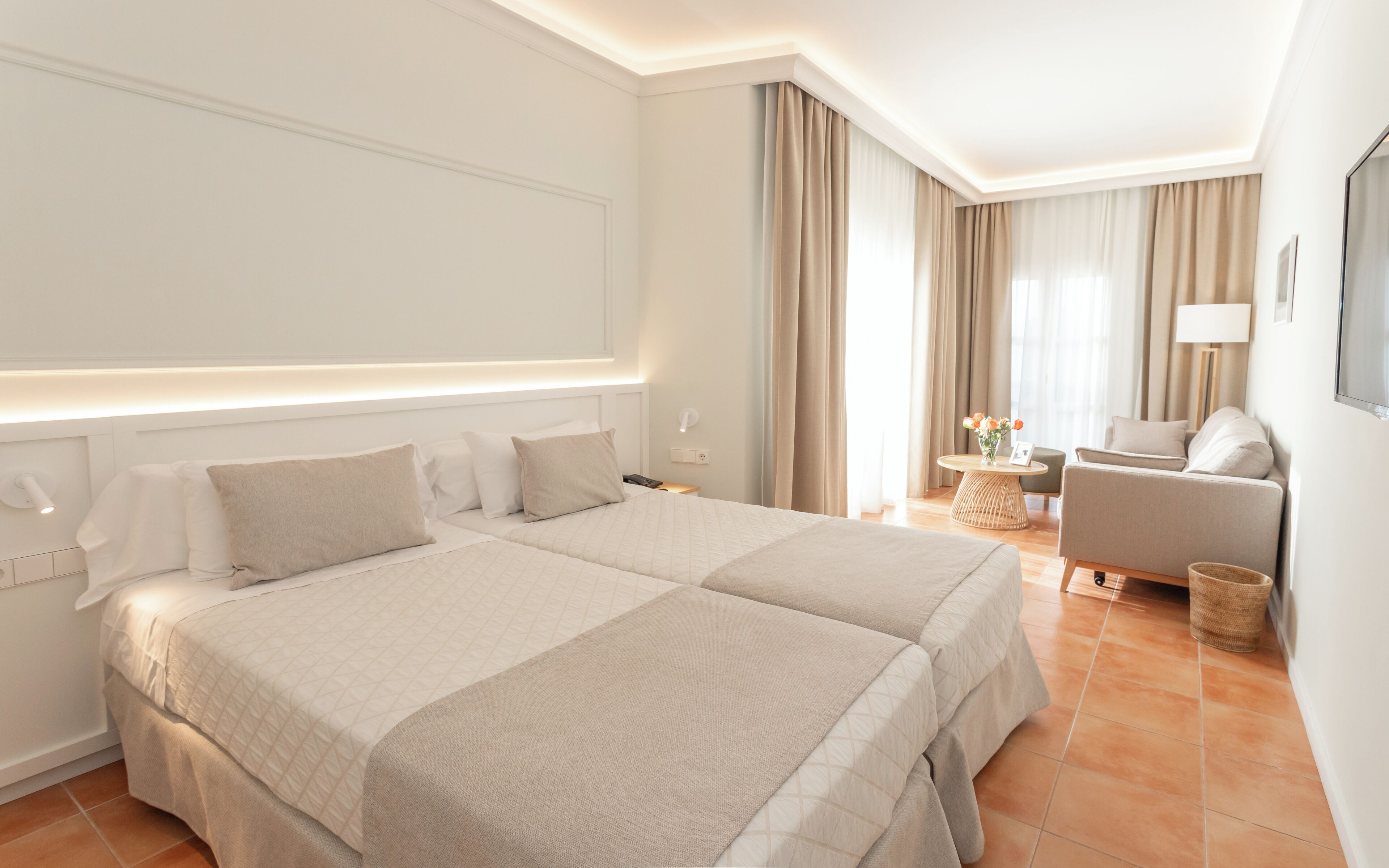 A hotel room with two beds, a sitting area with a chair and table, and a large window with curtains.