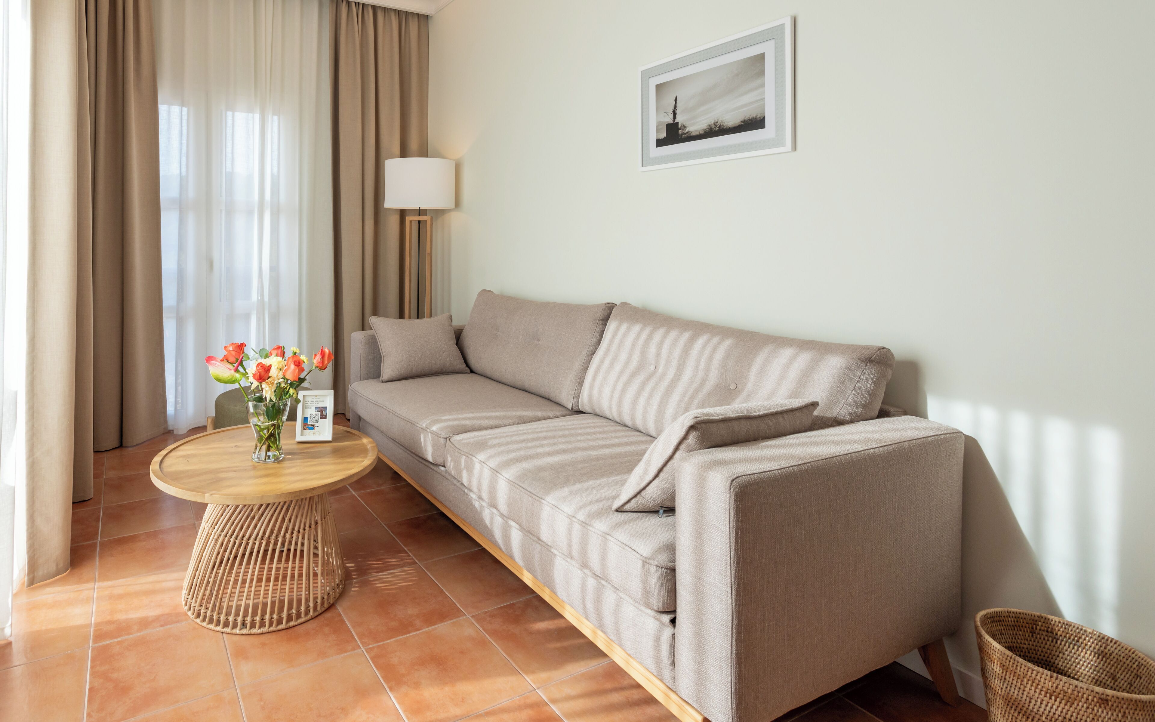 A modern living room with a beige sofa, a round wooden coffee table with a vase of flowers, and a lamp.