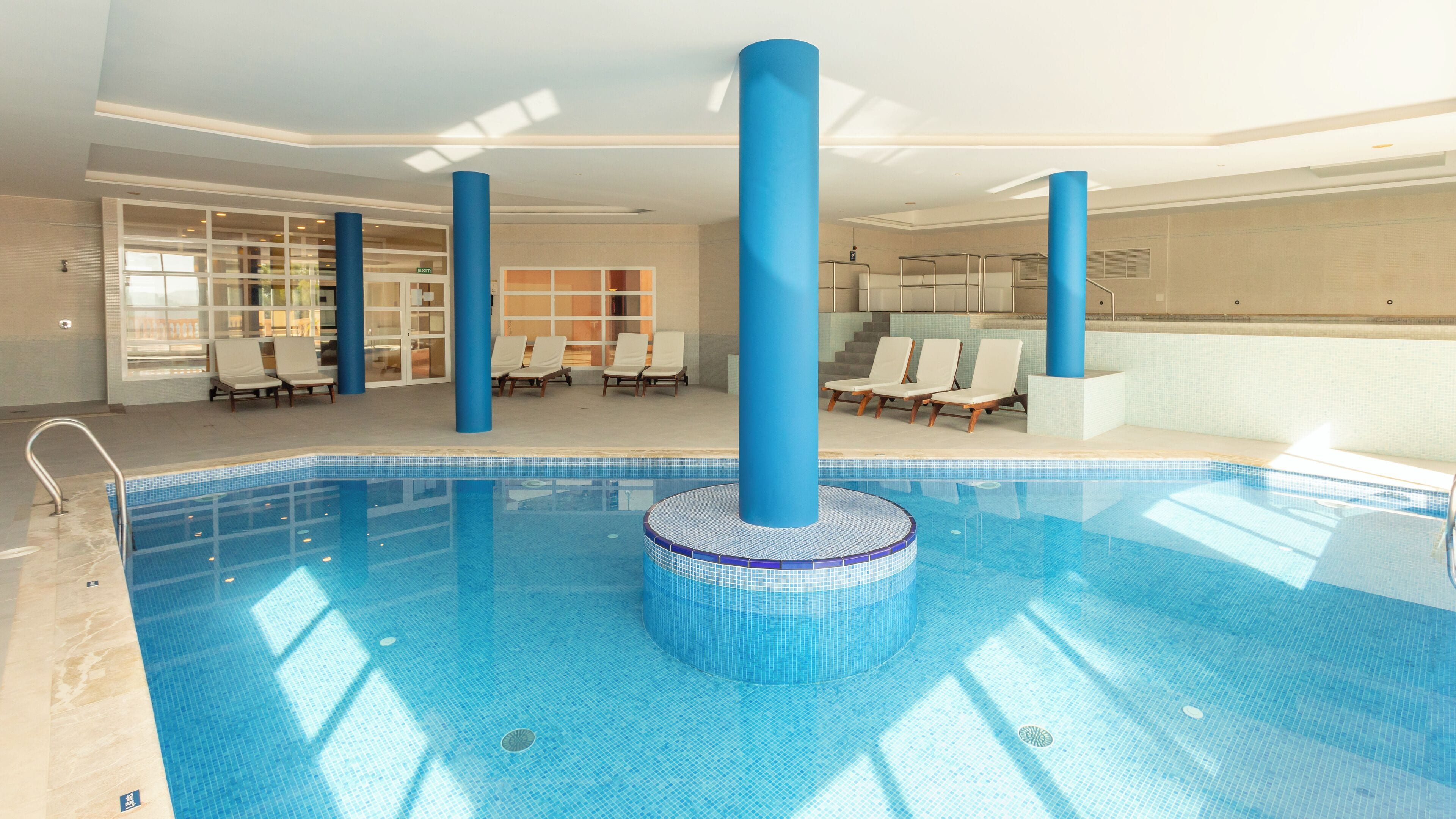 Ein Innenpool mit blauen Mosaikfliesen, umgeben von Liegestühlen und blauen Säulen.