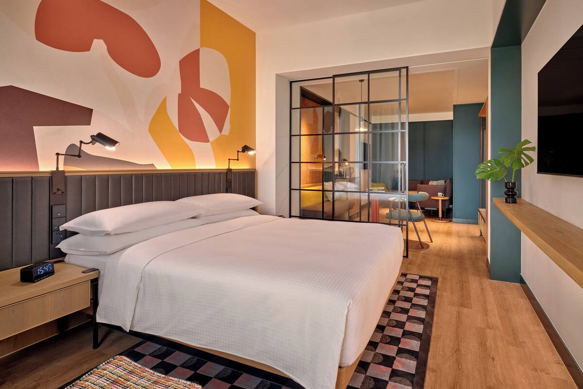 A modern hotel room with a large bed, a desk, a TV, and a view of the adjacent room.