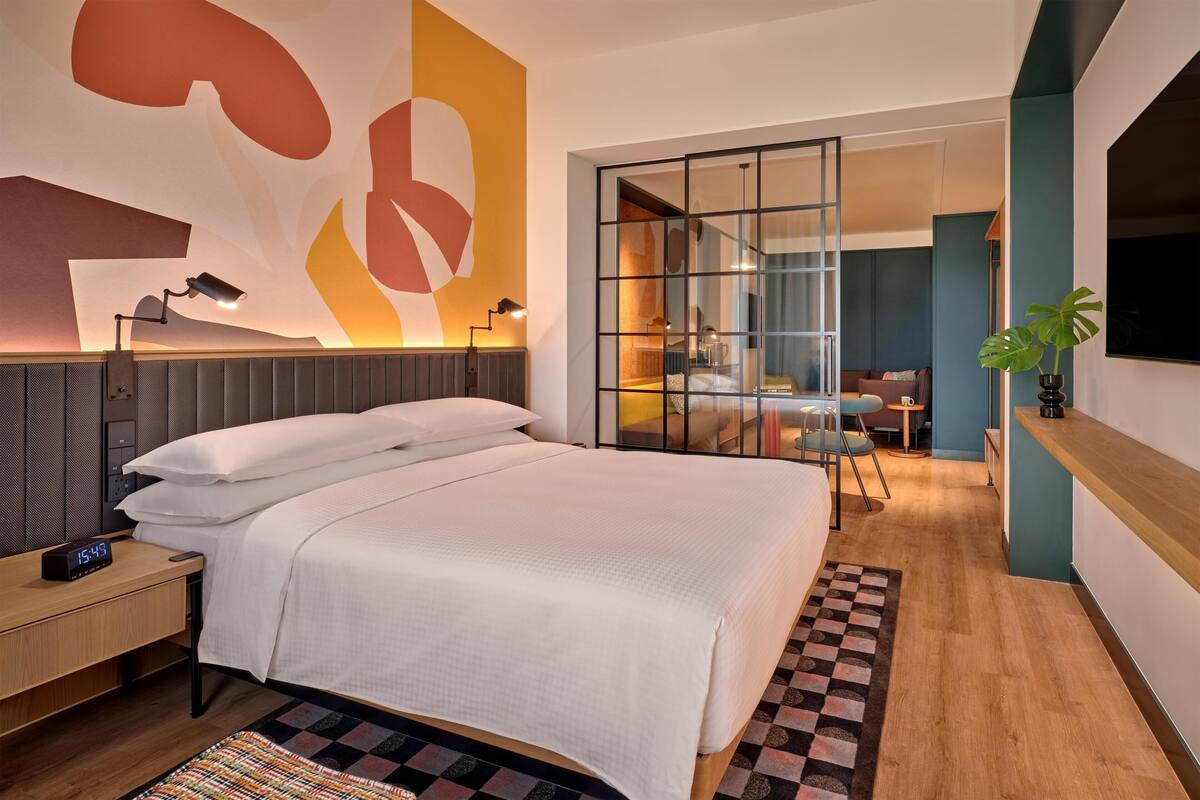 A modern hotel room with a large bed, a desk with a lamp, a TV, and a view into the dining area.