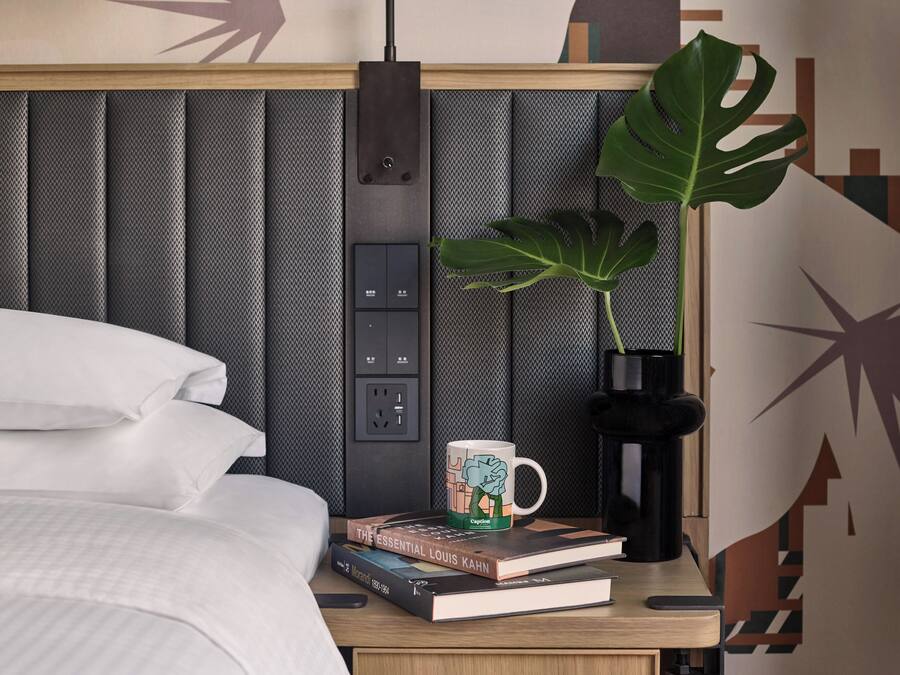 A neatly made bed with white linens, a bedside table with a book, a mug, and a plant.