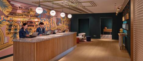 A reception desk with two staff members, a check-in counter, a wall with a colorful mural, and a modern ceiling design.