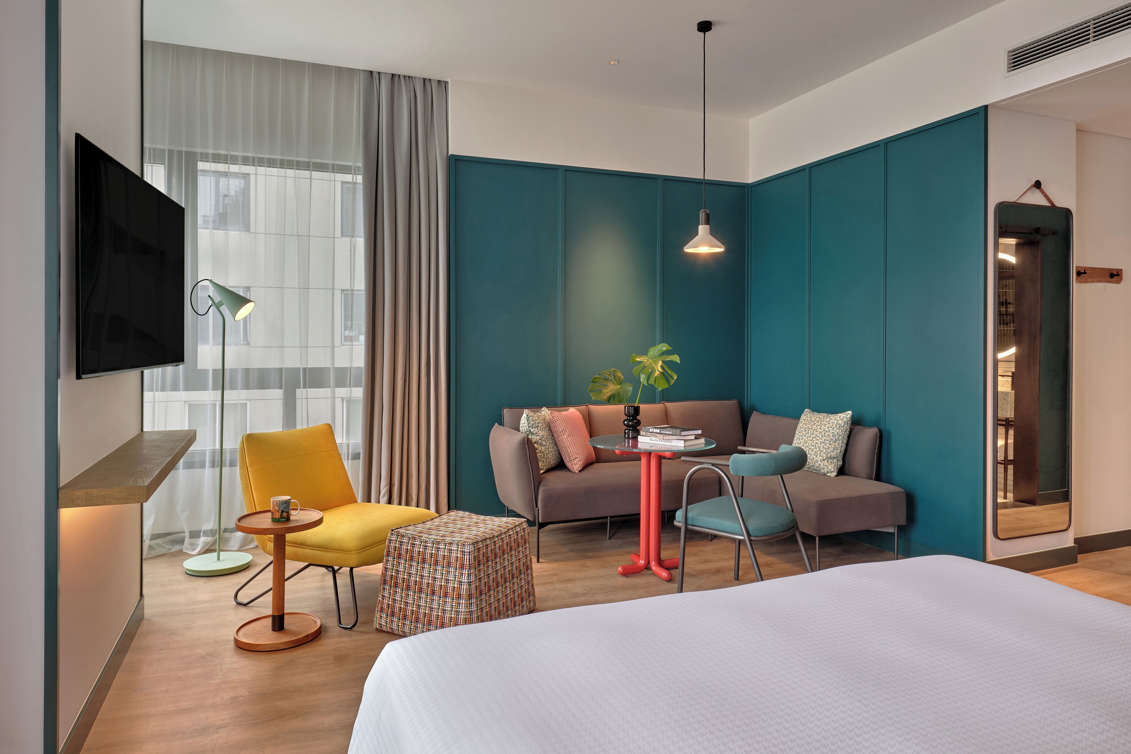 Une chambre d’hôtel moderne avec un grand lit, un canapé, un fauteuil jaune, une petite table et un téléviseur fixé au mur.