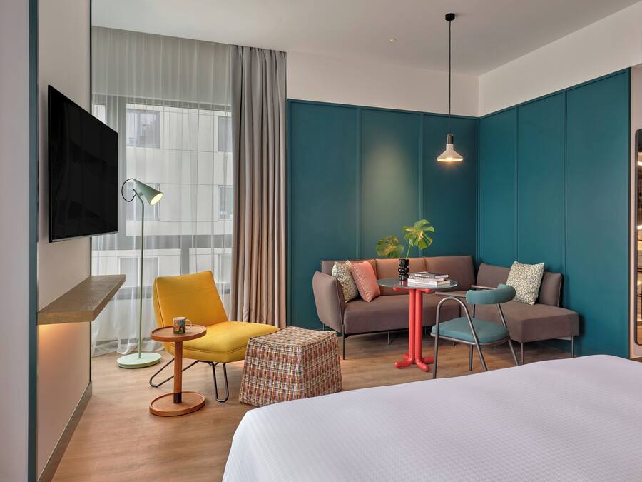 A modern hotel room with a large bed, a sofa, a yellow armchair, a small table, and a TV mounted on the wall.