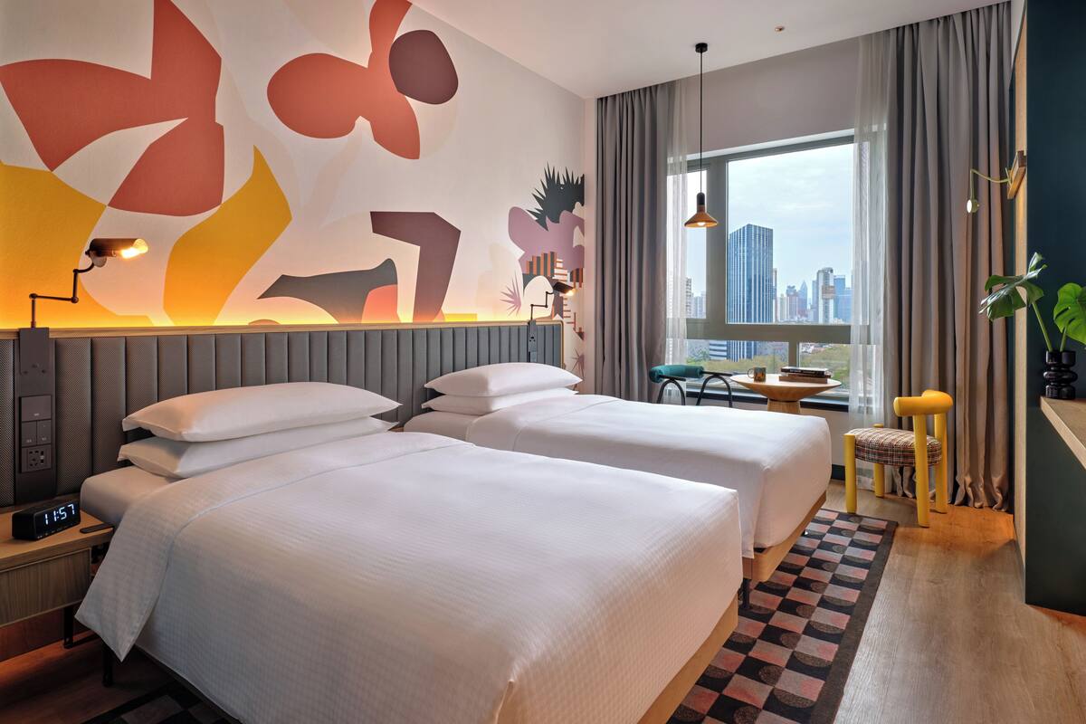 A hotel room with two beds, a desk, a chair, a window with a city view, and abstract wall art.