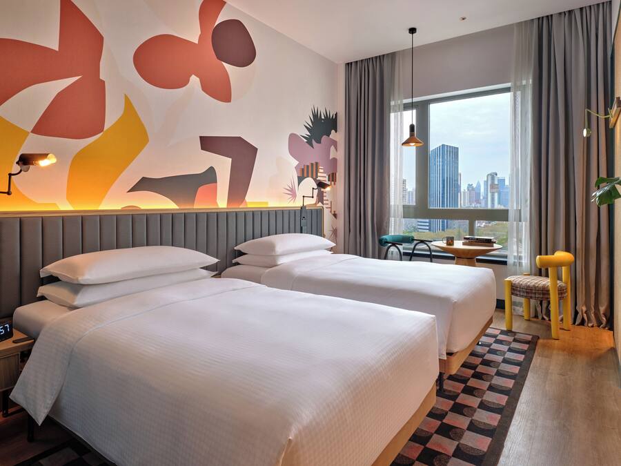 A hotel room with two beds, a desk, a chair, a window with a city view, and abstract wall art.