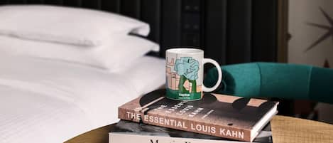 A book titled 'The Essential Louis Kahn' on a table, a mug with a caption, and a bed with white linens in the background.
