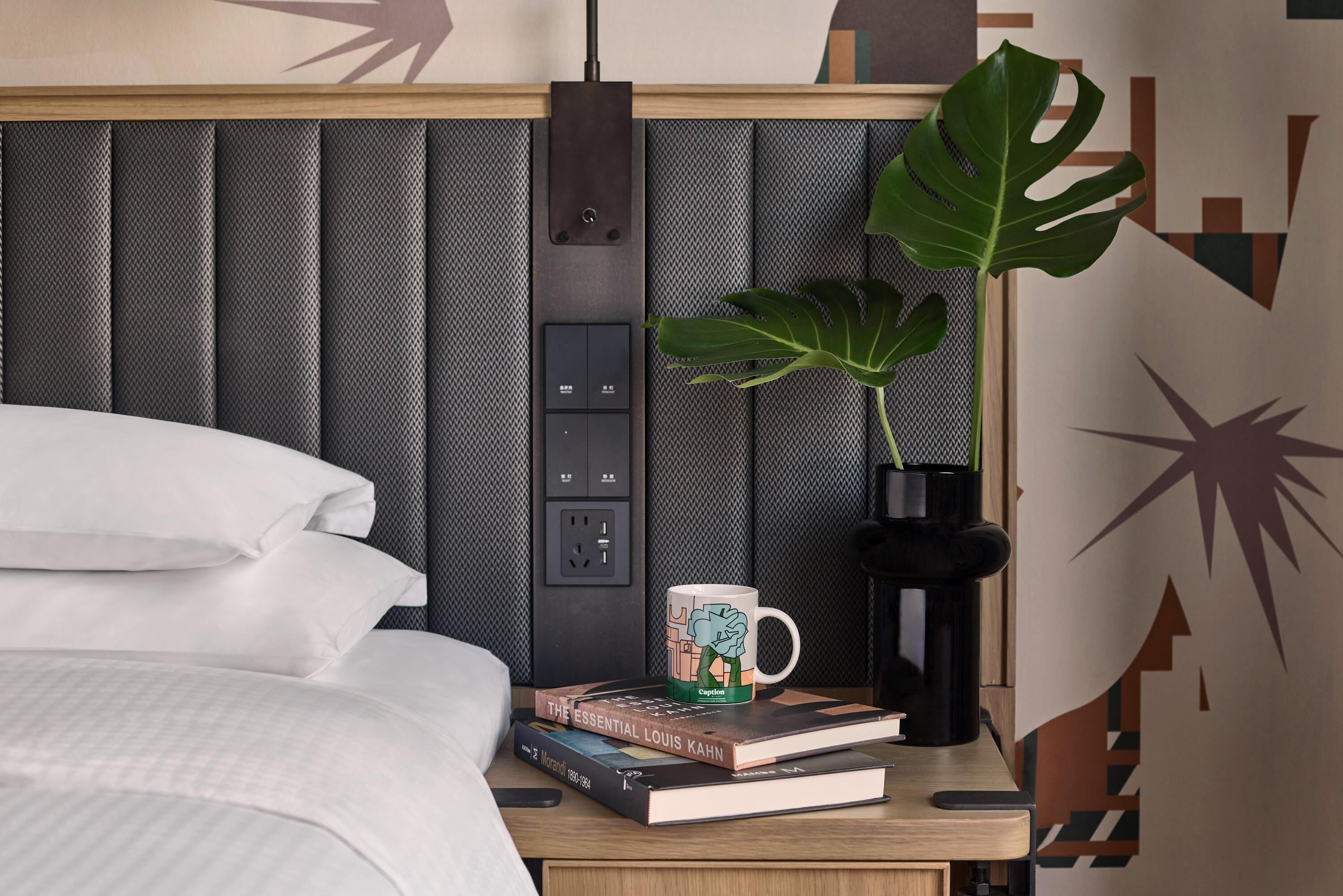 Una cama bien hecha con sábanas blancas, una mesita de noche con una taza, libros y una planta.