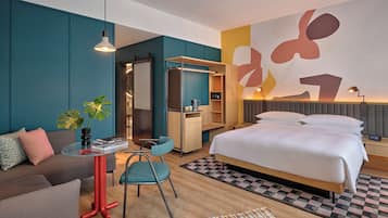 Moderni hotellihuone, jossa on sänky, työpöytä, tuoli, sohva, vaatekaappi ja suuri, abstrakti taideteos seinällä.