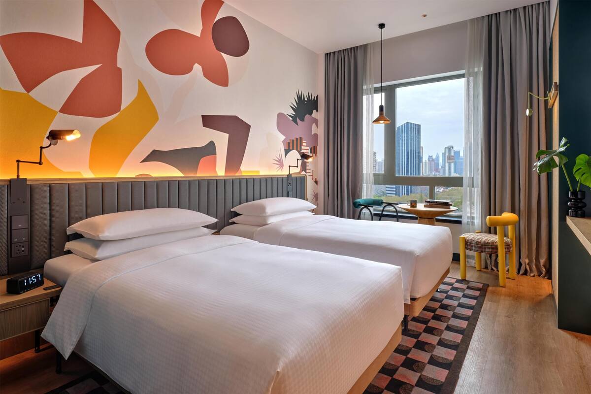 A hotel room with two beds, a desk, a chair, a window with a city view, and abstract wall art.