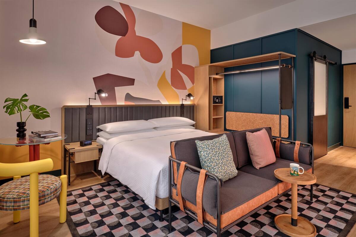 A modern hotel room with a bed, sofa, bedside table, lamp, and a wall with abstract art.