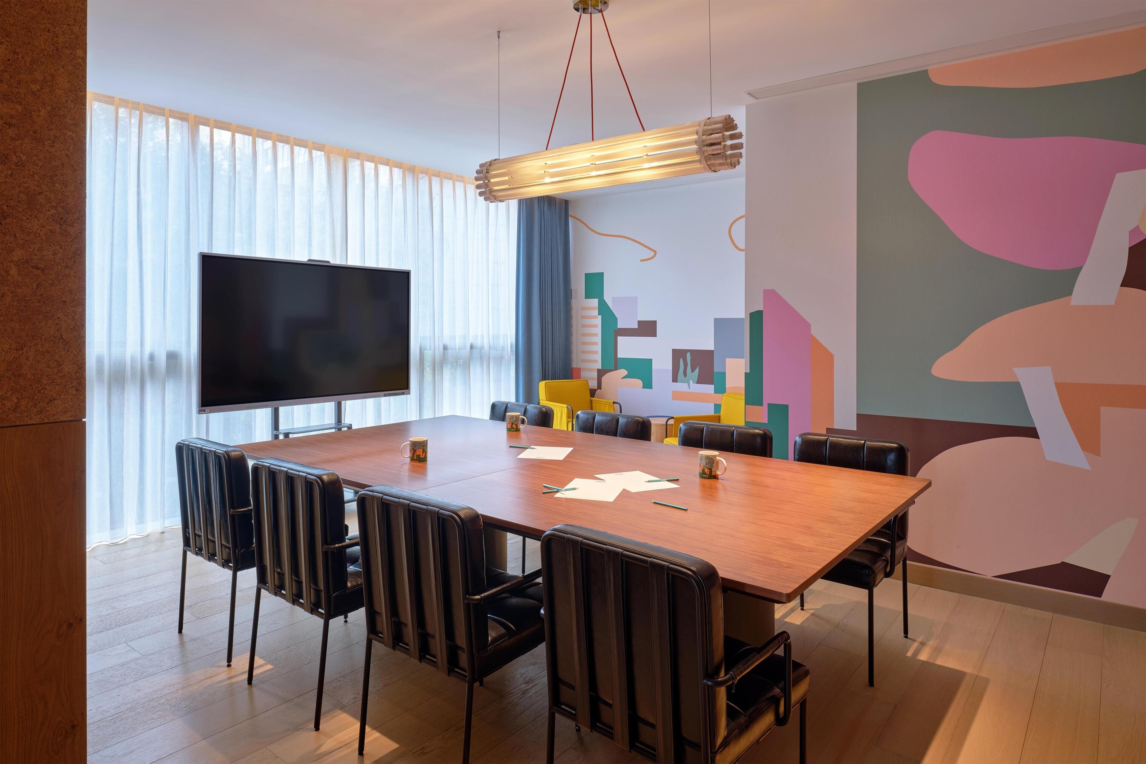 Een vergaderruimte met een grote tafel, stoelen, een flatscreen-tv en een kleurrijke abstracte muurschildering.