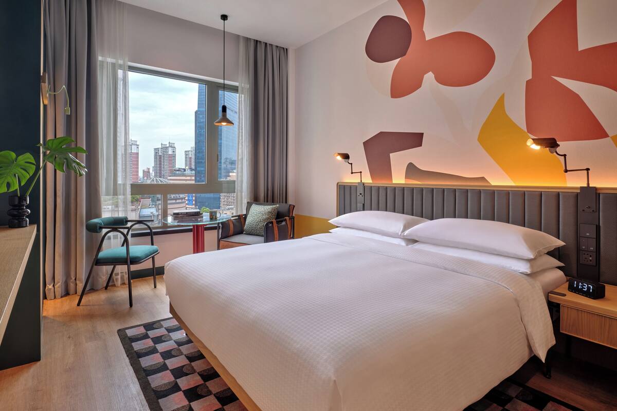 A hotel room with a large bed, a desk, a chair, a window with a city view, and abstract wall art.