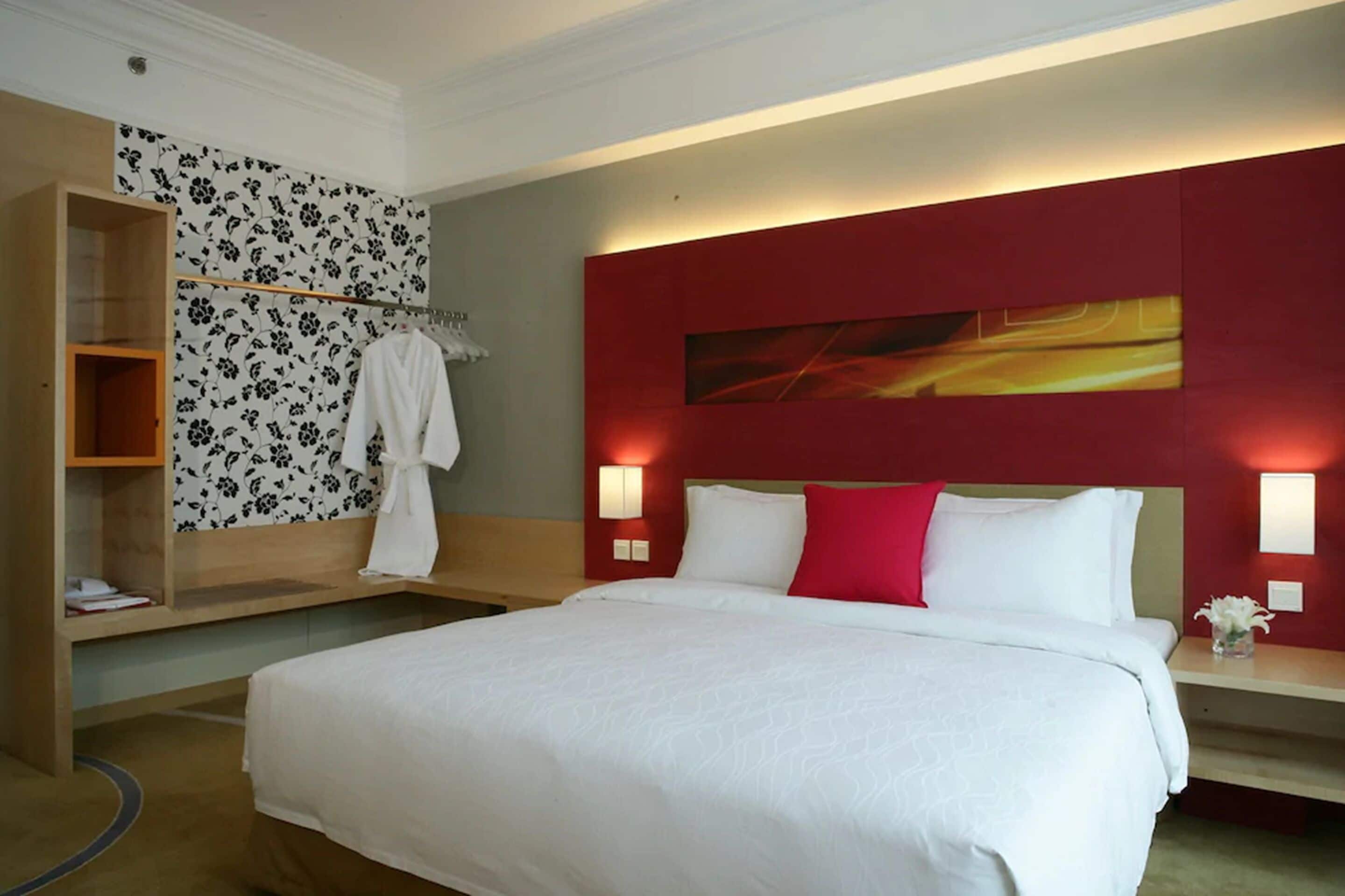 A hotel room with a large bed, a wooden wardrobe, a mirror, and a red accent wall with a painting.