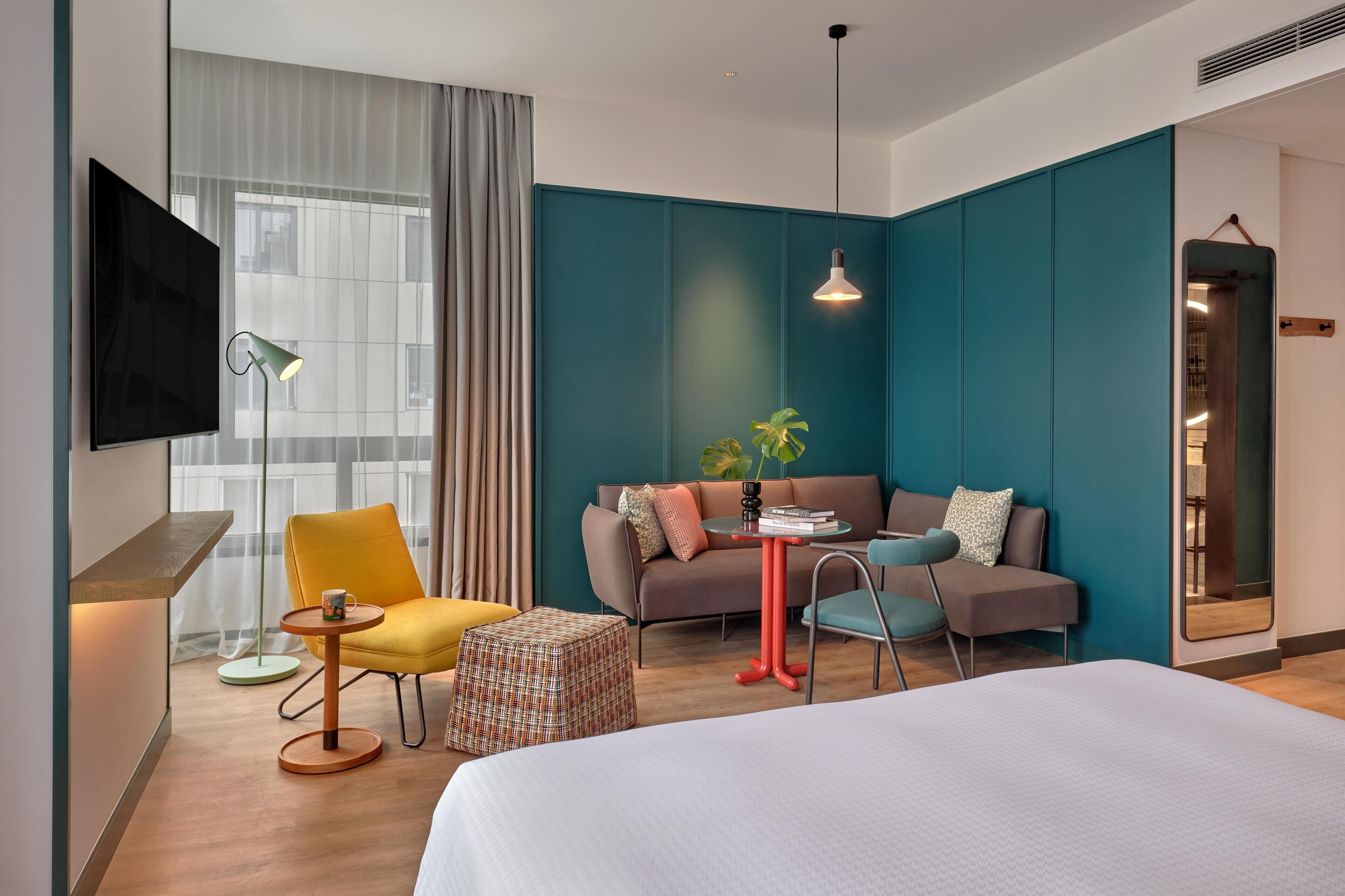 A modern hotel room with a large bed, a sofa, a yellow armchair, a small round table, a television, and a large window with curtains.