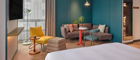 A modern hotel room with a large bed, a sofa, a yellow armchair, a small round table, a television, and a large window with curtains.