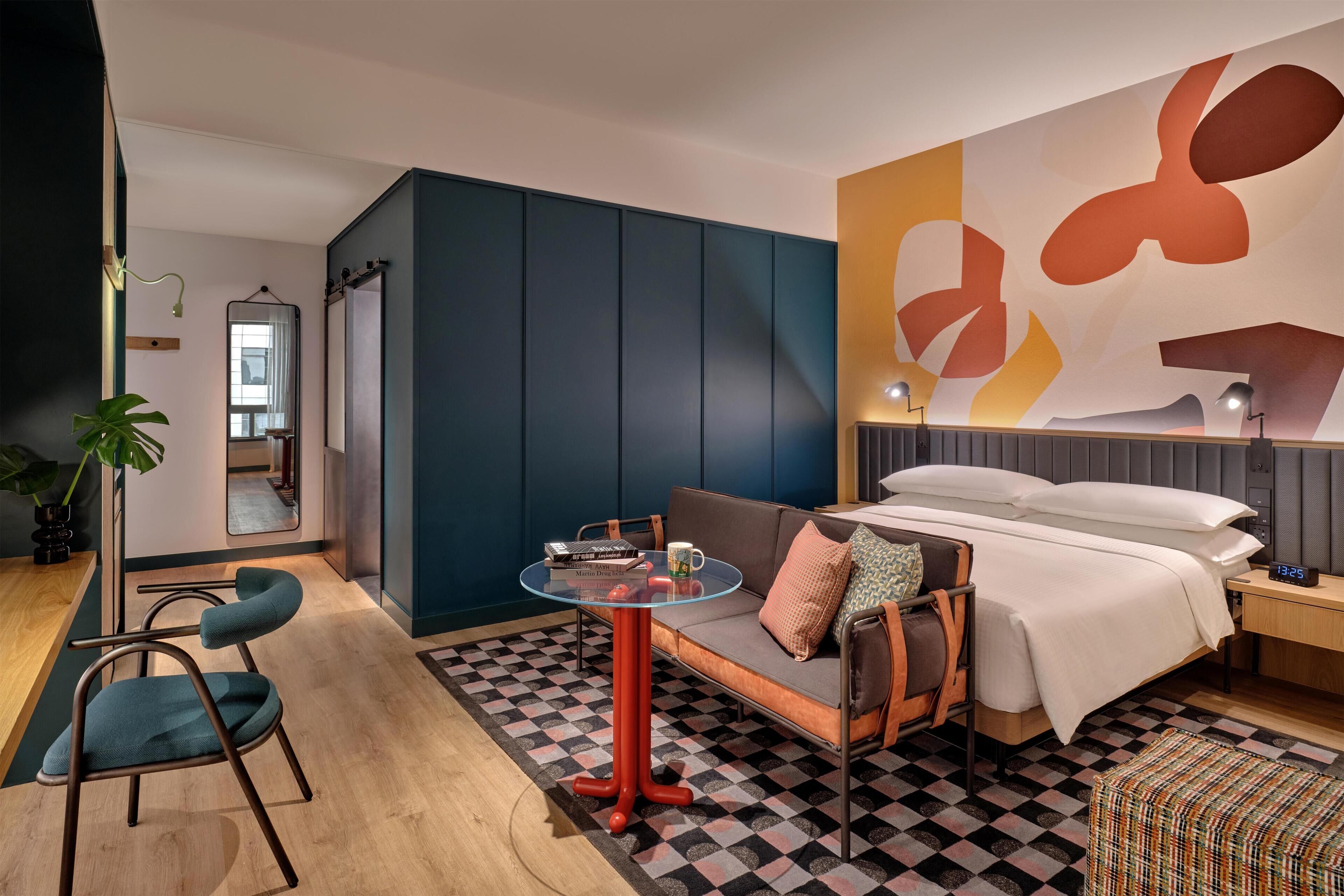 A modern hotel room with a sofa, a small round table, a bed, a nightstand, and a large abstract wall art piece.
