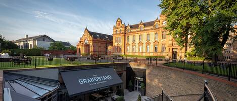 Un cortile con posti a sedere all'aperto al bar e ristorante The Grandstand, circondato da un edificio storico con una torre dell'orologio.