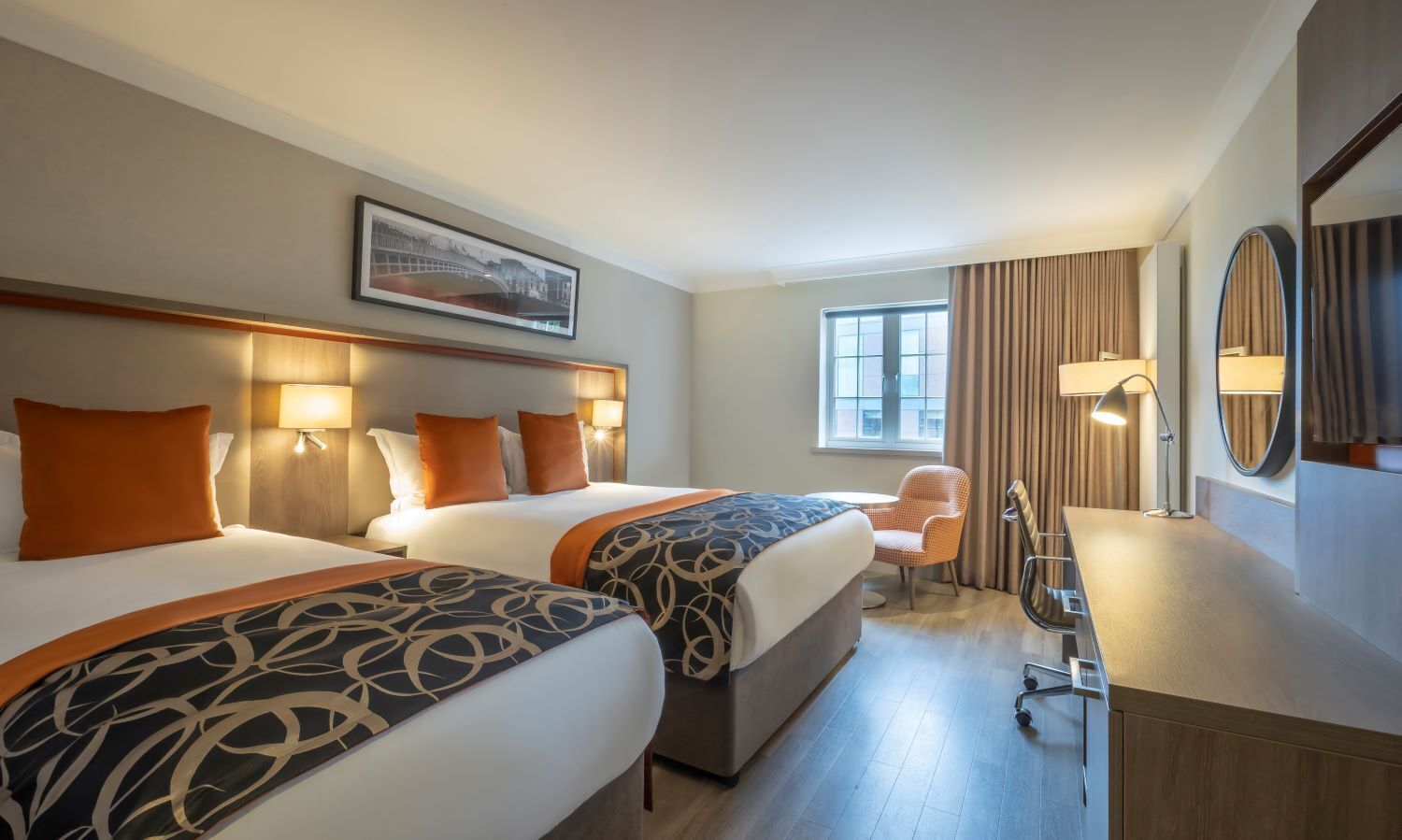 Habitación de hotel con dos camas, un escritorio, una silla, un ventanal grande y un cuadro en la pared.