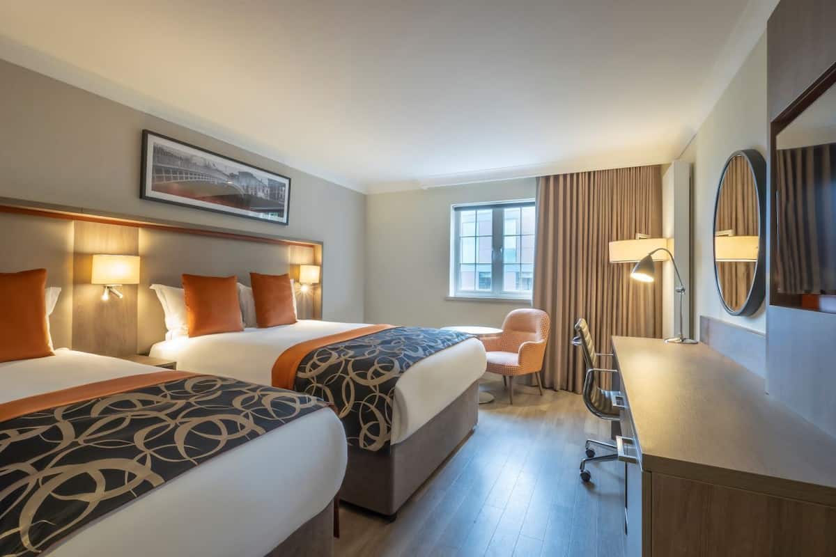 A hotel room with two beds, a desk, a chair, a large window, and a painting on the wall.