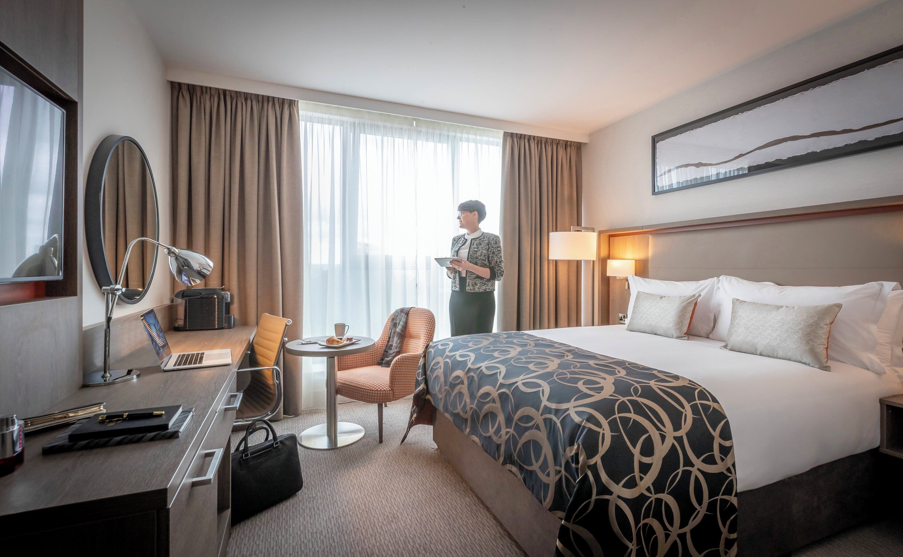 Une chambre d’hôtel avec un lit, un bureau, une chaise et une personne debout près de la fenêtre.