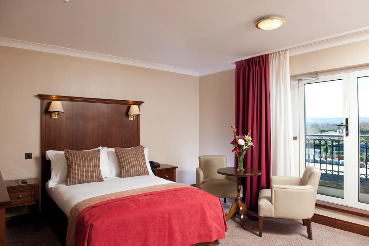 A hotel room with a bed, a nightstand, a chair, a small table with a vase, and a large window with a view.