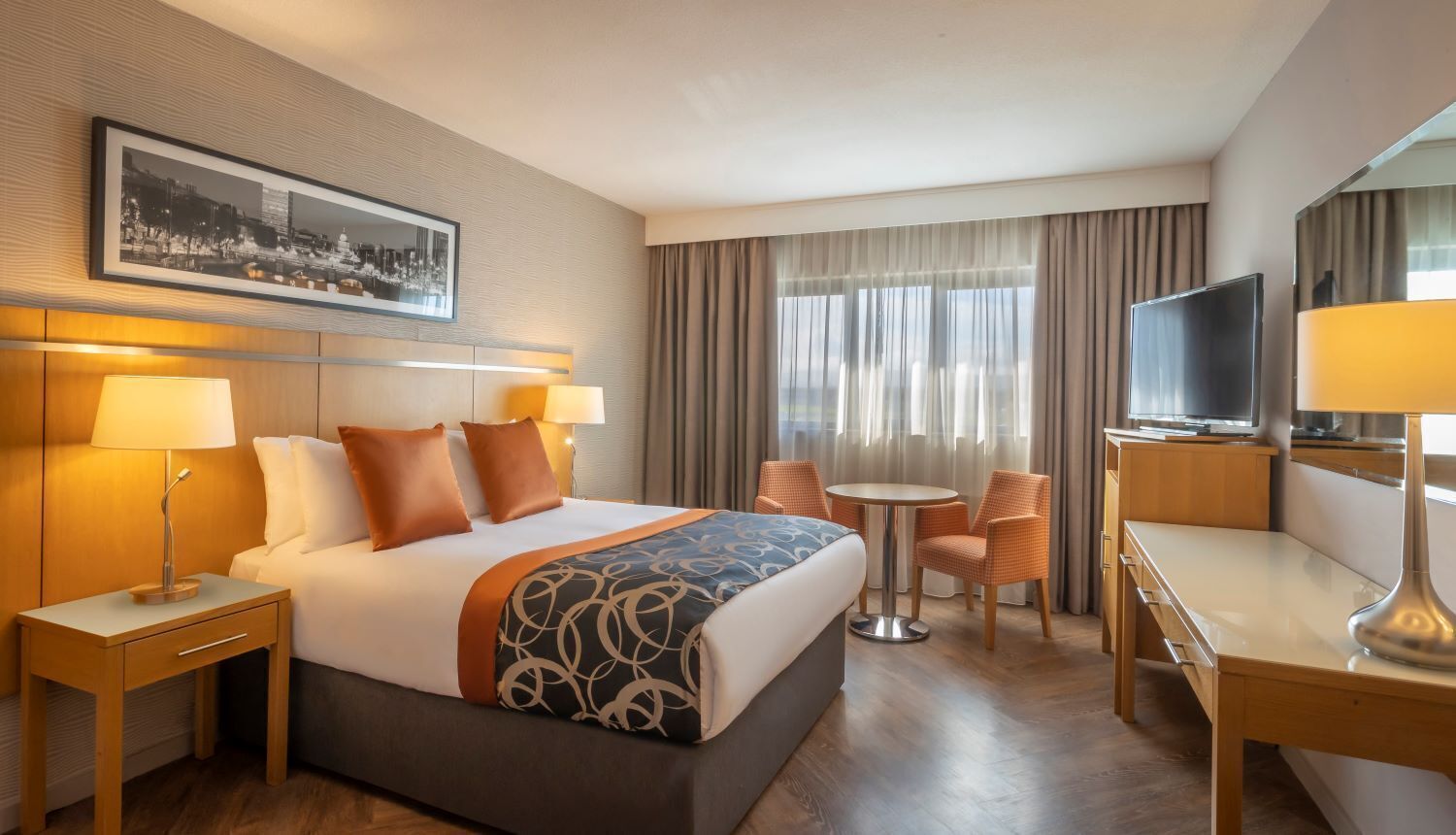 Habitación de hotel con cama, dos mesitas de noche, un escritorio, una silla, televisión y ventana con cortinas.