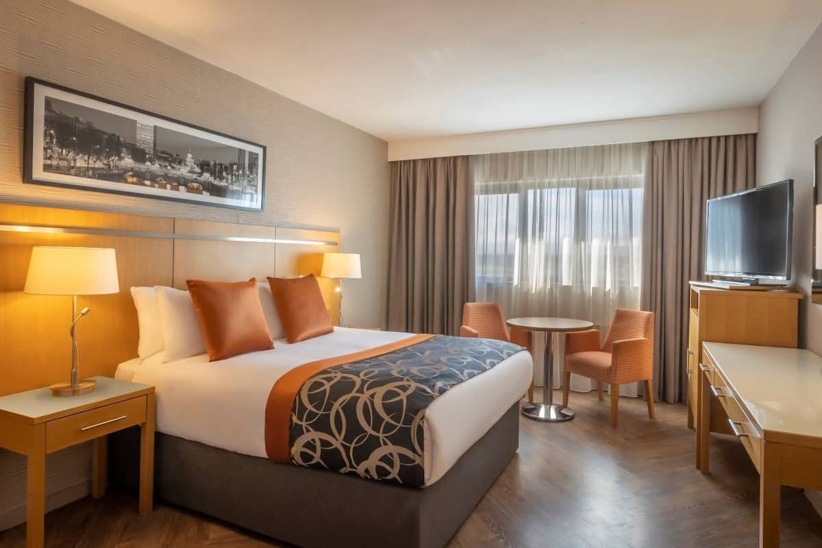 Habitación de hotel con cama, dos mesitas de noche, un escritorio, una silla, televisión y ventana con cortinas.
