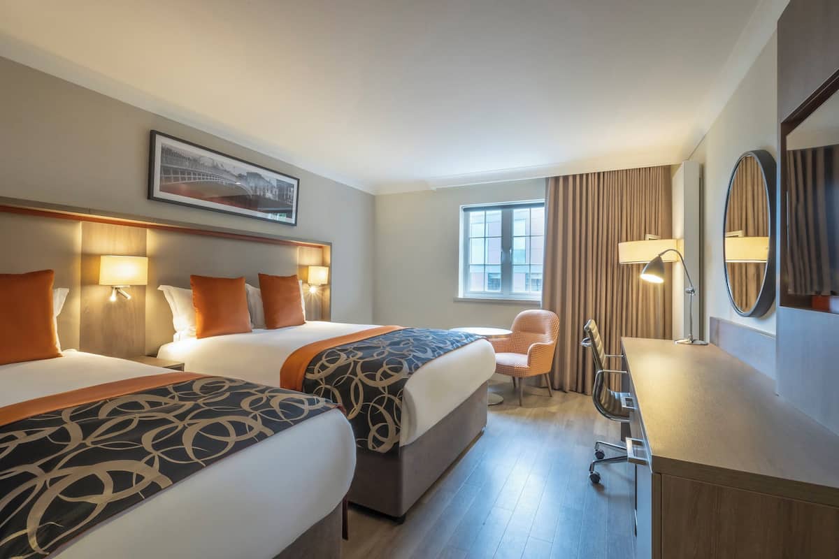 A hotel room with two beds, a desk, a chair, a window, and a painting on the wall.