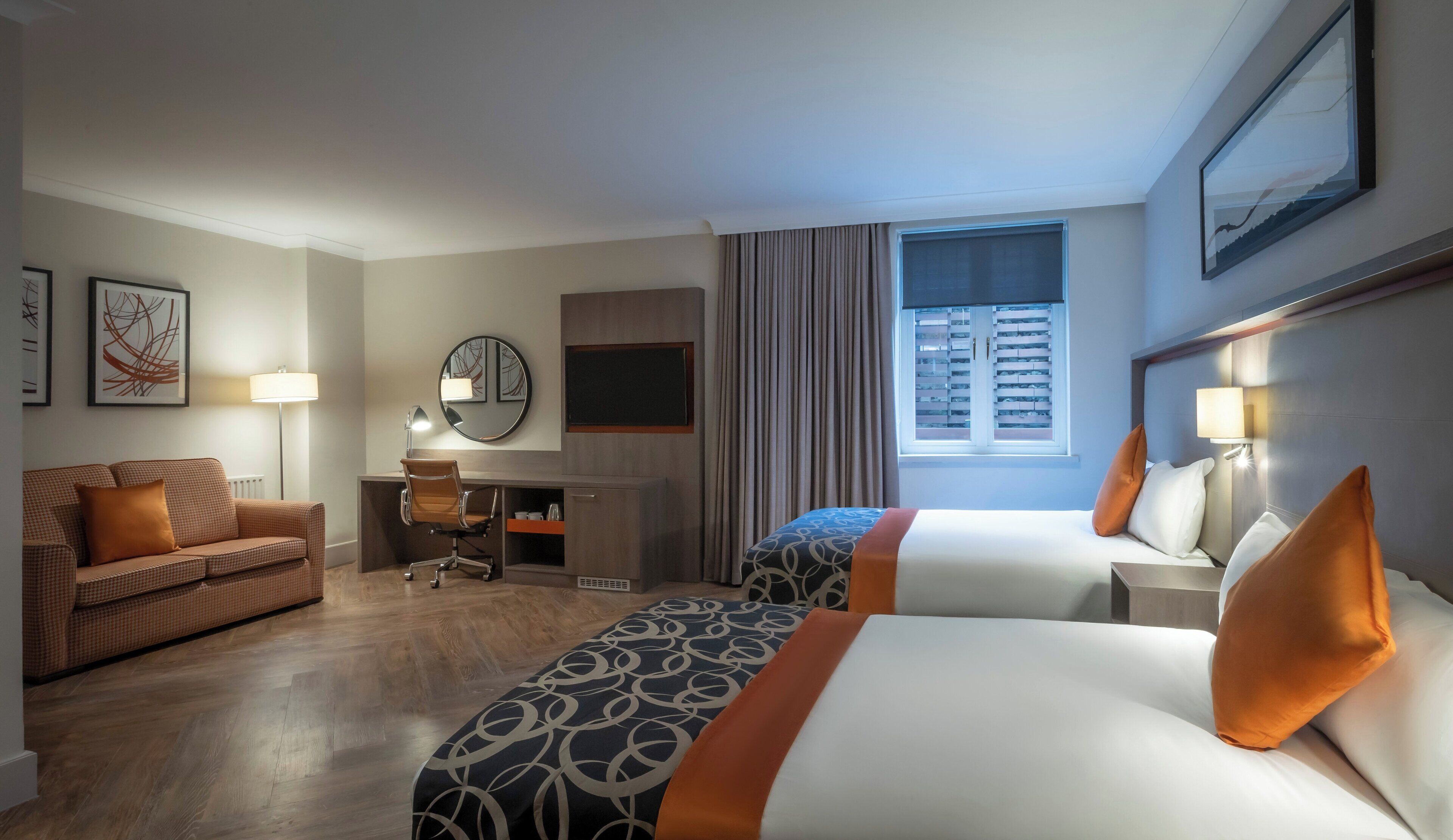 A hotel room with two beds, a desk, a chair, a couch, and a window with blinds.