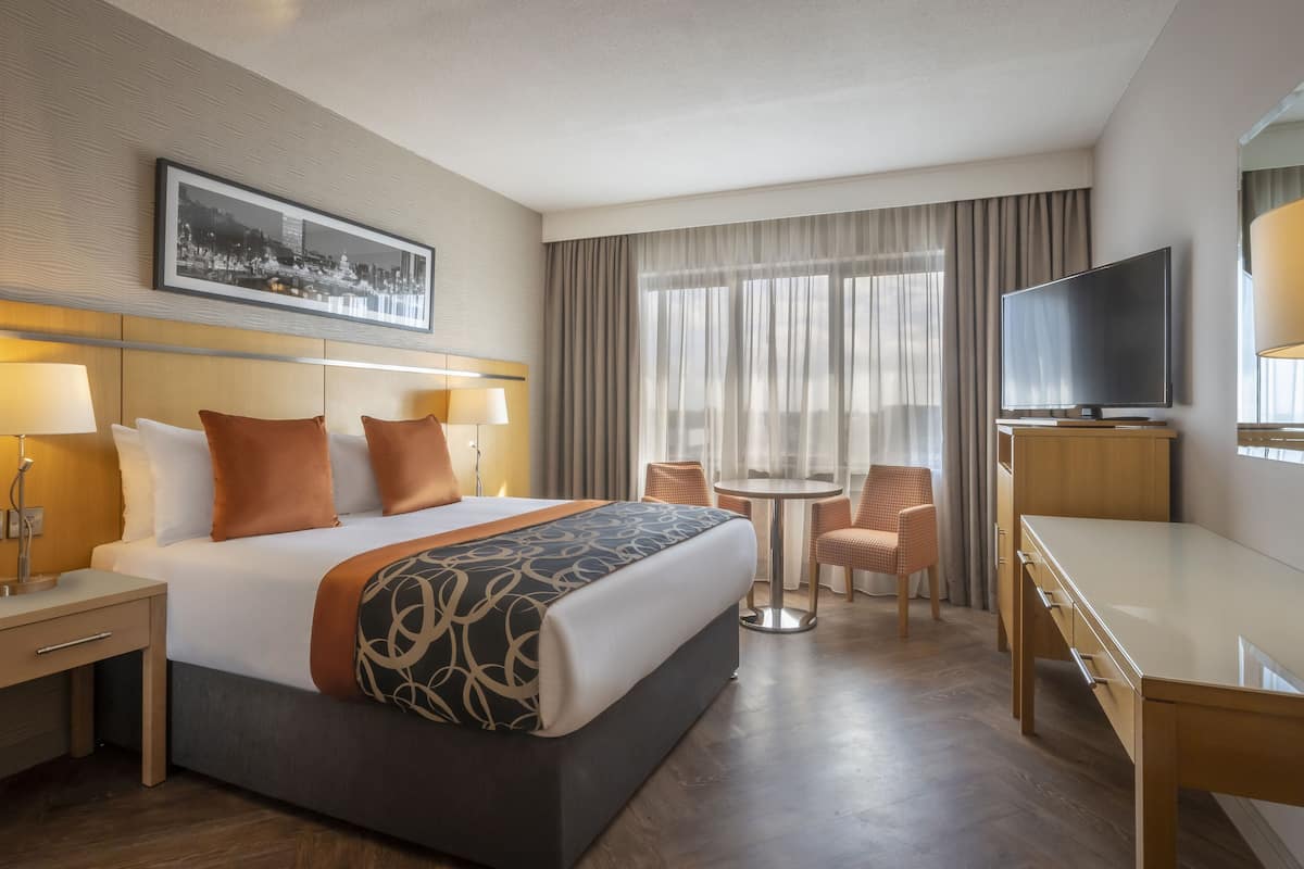 A hotel room with a bed, bedside tables, a desk, a chair, a TV, and a window with curtains.