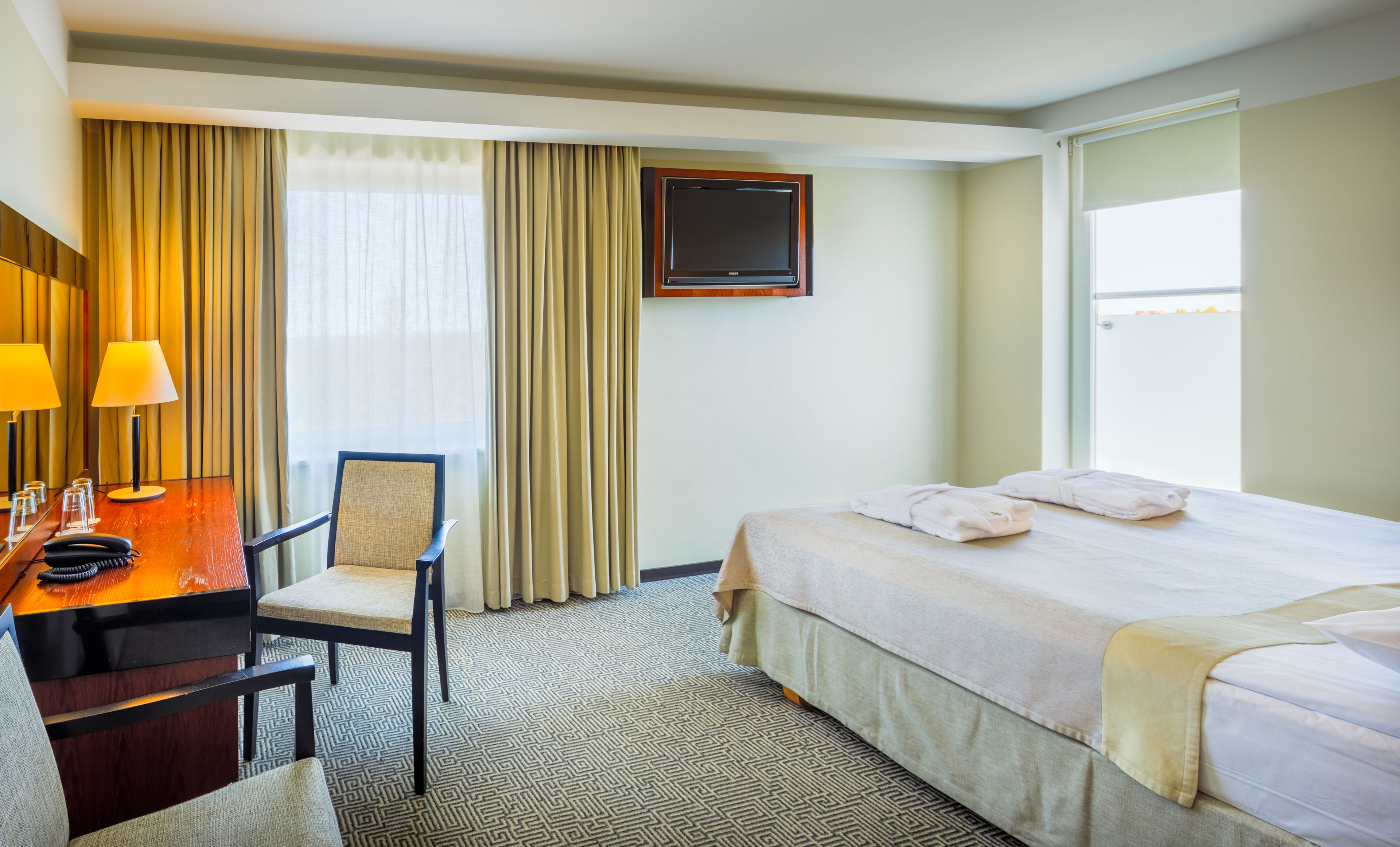 A hotel room with a bed, a desk with a telephone, a chair, a TV mounted on the wall, and a window with curtains.