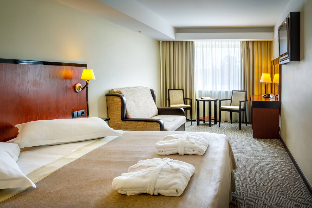 Quarto de hotel com uma cama grande, duas cadeiras, uma mesa pequena e uma escrivaninha com abajur.