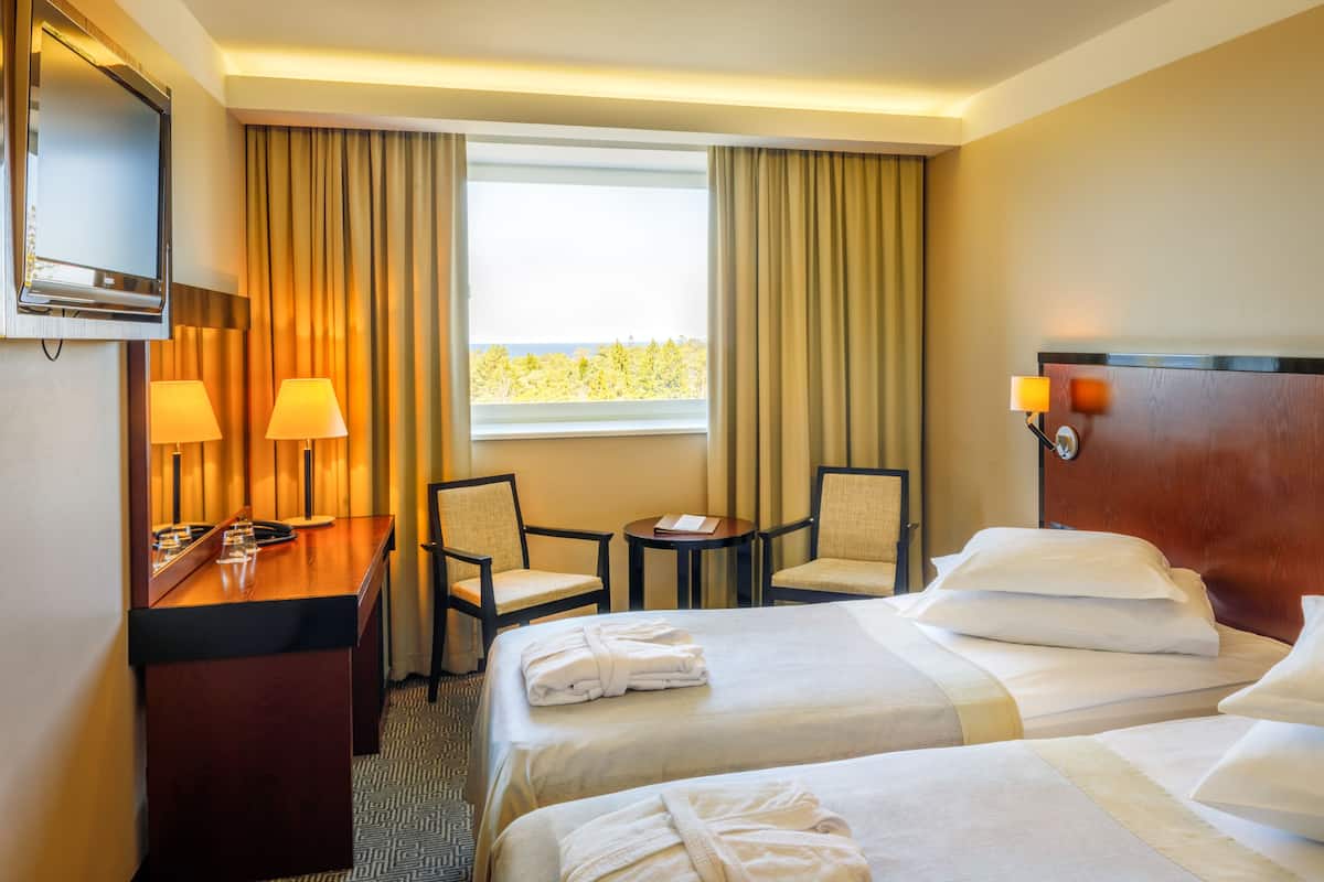 Quarto de hotel com duas camas, uma escrivaninha, duas cadeiras, uma mesinha, televisão e janela com cortinas.