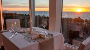 Une table dressée pour deux personnes, agrémentée d’un chandelier, placée près d’une grande fenêtre donnant sur un coucher de soleil sur la mer.